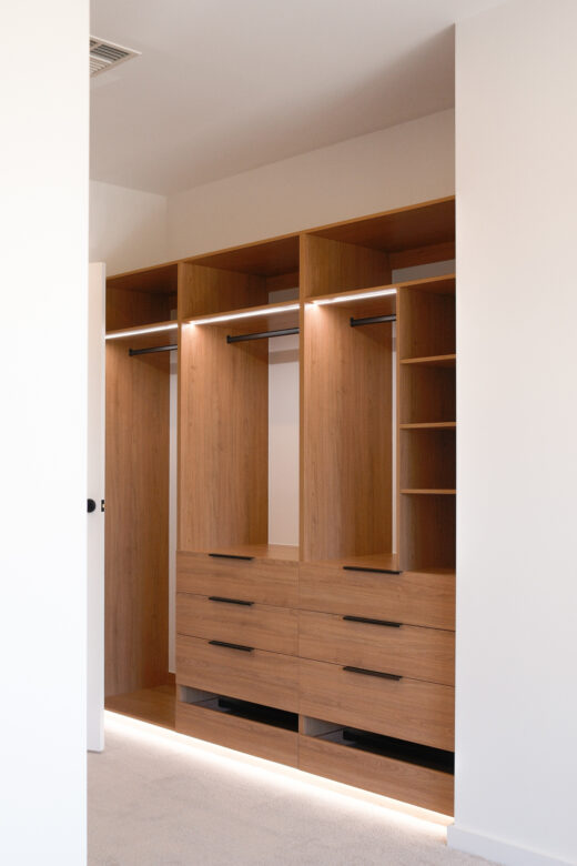 Custom-built semi-carcass walk-in robe in Polytec Tasmanian Oak featuring Artia Jay black lip handles and integrated Häfele LED lighting, by Packers.