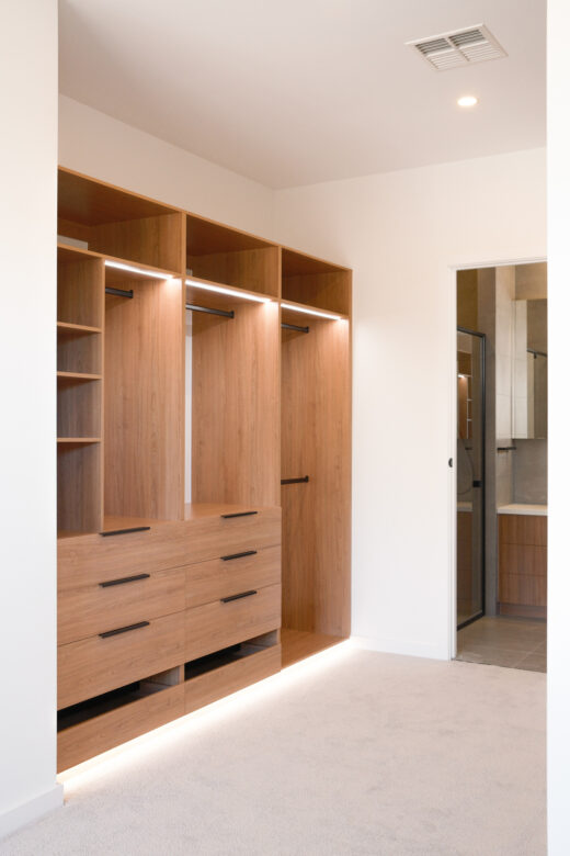 Custom-built semi-carcass walk-in robe in Polytec Tasmanian Oak featuring Artia Jay black lip handles and integrated Häfele LED lighting, by Packers.